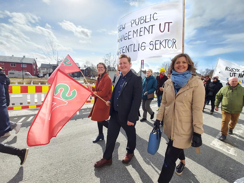 Bilde fra 1.mai tog. Tre personer fra SV i front. To av dem holder et flagg med SV sin logo. De går foran en fane hvor det står New public management ut av offentlig sektor. Bilde.